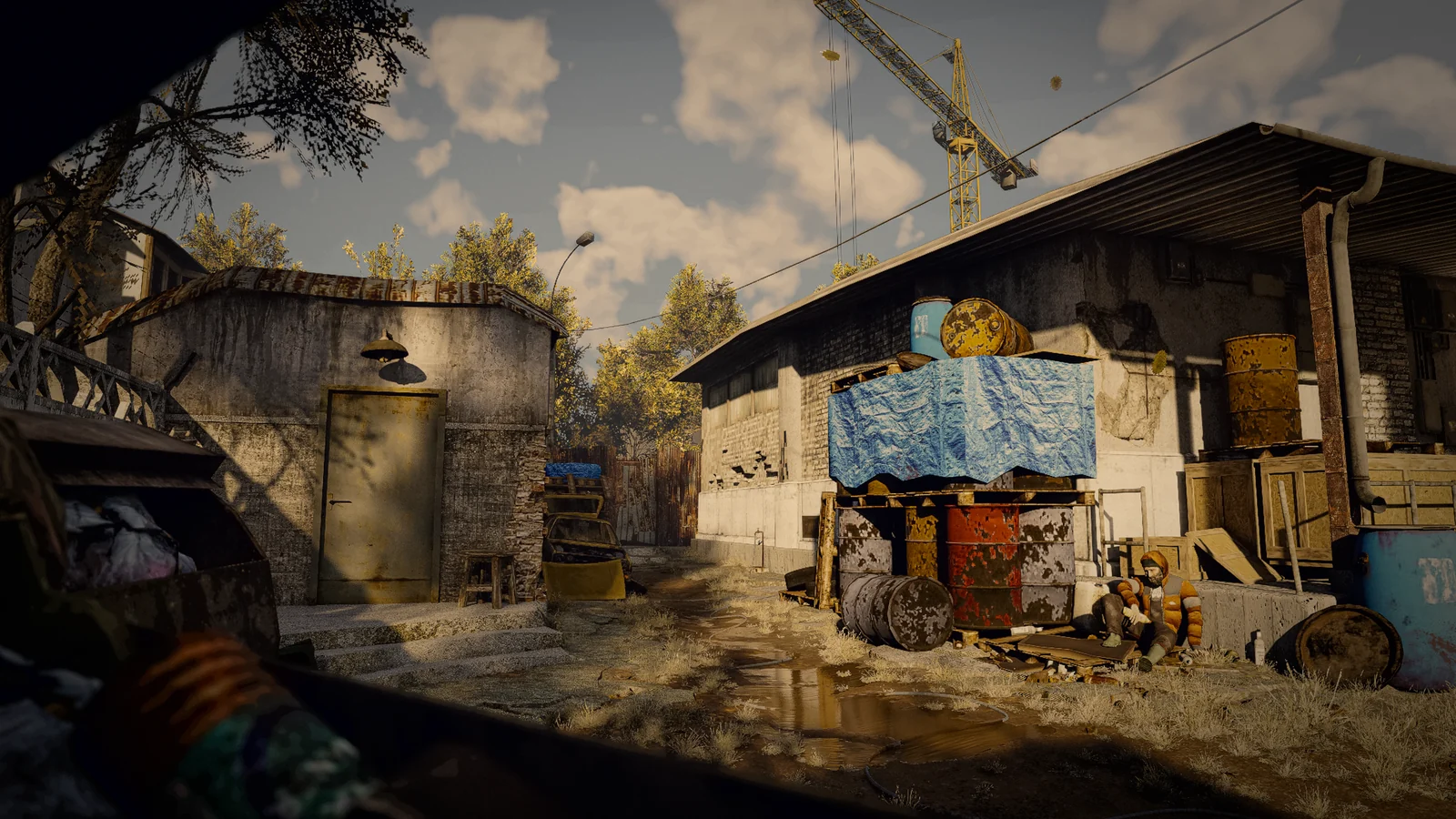 An industrial yard with stacked barrels, a blue tarpaulin and a man sitting by a warehouse.
